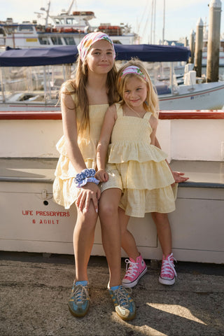 Two young girls smile at the camera on a dock in front of boats, both wearing matching Roller Rabbit Kids Petal Eyelet Kenia Dresses. The older girl sports sneakers and a blue scrunchie, while the younger wears pink shoes.