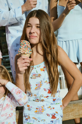 Young child with long brown hair stands barefoot, wearing the Roller Rabbit Kids Spain Monkey Noa Set—light blue Pima cotton loungewear with playful monkey prints and blue trim—against a plain white background.