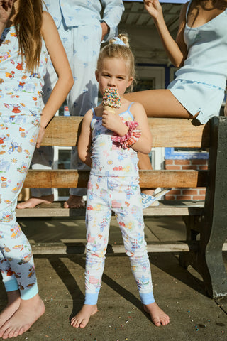 A young girl in pastel Roller Rabbit Kids Italy Monkey Noa Set pajamas stands barefoot on a wooden bench, eating a sprinkle-covered ice cream cone as sunlight highlights the playful scene with two adults and another child partially visible.