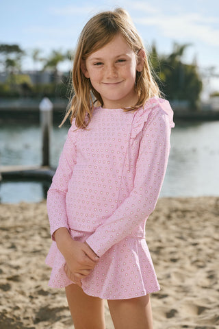 A young girl with light brown hair stands on a sandy beach, smiling in the Roller Rabbit Kids Hearts Ruffle Rashguard. The long-sleeve rashguard features a heart print. Water, trees, and a dock are visible in the background.