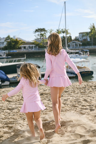Two young girls in matching pink outfits walking on a sandy beach with boats and houses in the background.