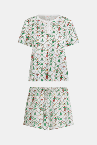 A woman wears a SERFLEX Deck The Halls Bailey Pajamas set featuring a short-sleeved top and shorts with a white background and a repeating Christmas-themed pattern of elves, snowmen, and evergreen trees.