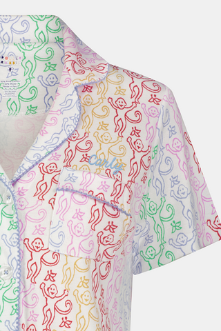 A woman with curly hair, standing and smiling slightly, wears Roller Rabbit’s Disco Monkey Wyatt Polo Pajamas: a short-sleeve button-up top and matching ruffled shorts featuring a pastel multicolor abstract pattern on white.