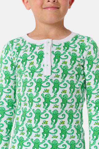 A young boy smiles at the camera, barefoot in Roller Rabbit’s Kids Monkey Pajamas— a matching top and bottom set featuring playful monkey prints and white cuffs, made from 100 pima cotton, against a plain white background.