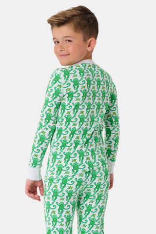 A young boy smiles at the camera, barefoot in Roller Rabbit’s Kids Monkey Pajamas— a matching top and bottom set featuring playful monkey prints and white cuffs, made from 100 pima cotton, against a plain white background.