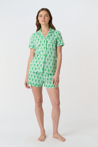 A woman stands barefoot on a white background wearing Roller Rabbit’s Monkey Polo Pajamas—a matching set with a short-sleeve button-up top and shorts featuring a playful green and white pattern.