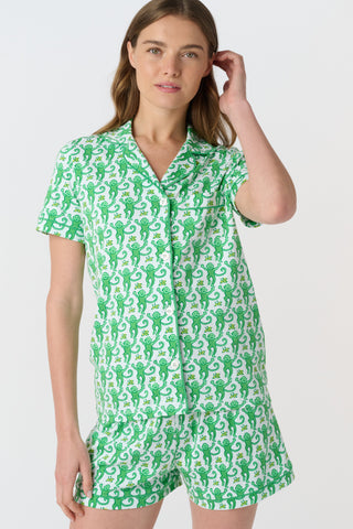 A woman stands barefoot on a white background wearing Roller Rabbit’s Monkey Polo Pajamas—a matching set with a short-sleeve button-up top and shorts featuring a playful green and white pattern.