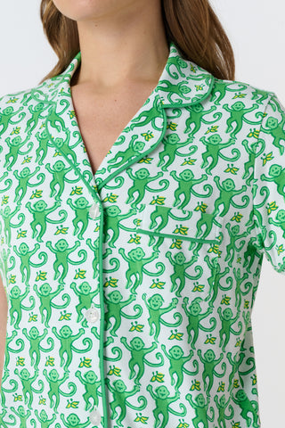 A woman stands barefoot on a white background wearing Roller Rabbit’s Monkey Polo Pajamas—a matching set with a short-sleeve button-up top and shorts featuring a playful green and white pattern.