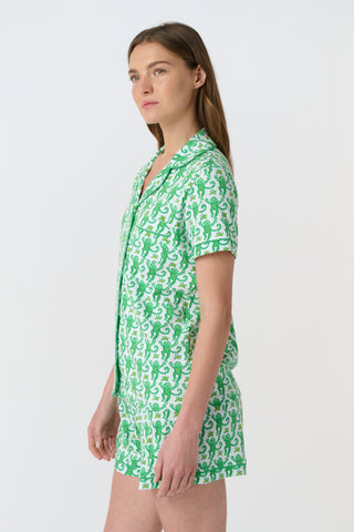 A woman stands barefoot on a white background wearing Roller Rabbit’s Monkey Polo Pajamas—a matching set with a short-sleeve button-up top and shorts featuring a playful green and white pattern.