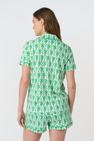A woman stands barefoot on a white background wearing Roller Rabbit’s Monkey Polo Pajamas—a matching set with a short-sleeve button-up top and shorts featuring a playful green and white pattern.