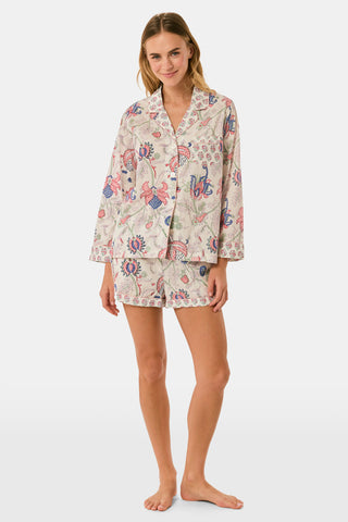 A woman stands against a plain white background, smiling slightly as she models the Amanda Leila Set from Roller Rabbit—a vintage-inspired cotton loungewear ensemble featuring a long-sleeve, button-up pajama top and matching shorts adorned with a colorful floral pattern.
