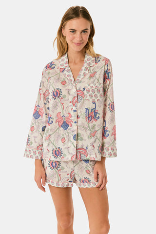 A woman stands against a plain white background, smiling slightly as she models the Amanda Leila Set from Roller Rabbit—a vintage-inspired cotton loungewear ensemble featuring a long-sleeve, button-up pajama top and matching shorts adorned with a colorful floral pattern.