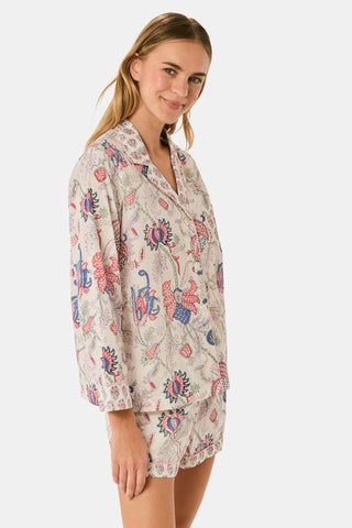 A woman stands against a plain white background, smiling slightly as she models the Amanda Leila Set from Roller Rabbit—a vintage-inspired cotton loungewear ensemble featuring a long-sleeve, button-up pajama top and matching shorts adorned with a colorful floral pattern.