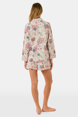 A woman stands against a plain white background, smiling slightly as she models the Amanda Leila Set from Roller Rabbit—a vintage-inspired cotton loungewear ensemble featuring a long-sleeve, button-up pajama top and matching shorts adorned with a colorful floral pattern.