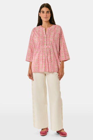 A woman models the Cora Floral Stripe Arles Top by Roller Rabbit—a loose, long-sleeved tunic in autumnal pink featuring a floral stripe block print and pintuck pleats on the chest—paired with white pants, against a plain light background.