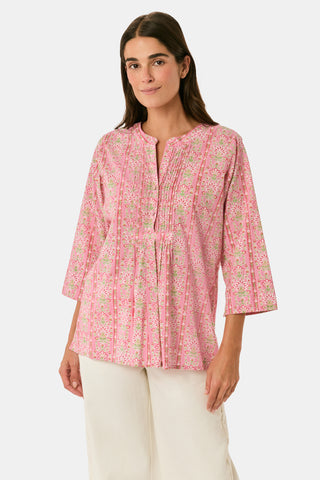 A woman models the Cora Floral Stripe Arles Top by Roller Rabbit—a loose, long-sleeved tunic in autumnal pink featuring a floral stripe block print and pintuck pleats on the chest—paired with white pants, against a plain light background.