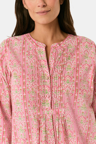 A woman models the Cora Floral Stripe Arles Top by Roller Rabbit—a loose, long-sleeved tunic in autumnal pink featuring a floral stripe block print and pintuck pleats on the chest—paired with white pants, against a plain light background.