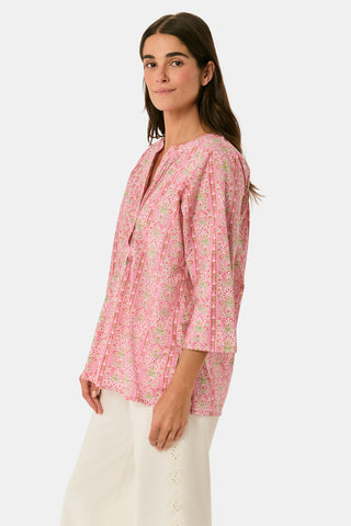 A woman models the Cora Floral Stripe Arles Top by Roller Rabbit—a loose, long-sleeved tunic in autumnal pink featuring a floral stripe block print and pintuck pleats on the chest—paired with white pants, against a plain light background.