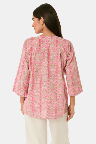 A woman models the Cora Floral Stripe Arles Top by Roller Rabbit—a loose, long-sleeved tunic in autumnal pink featuring a floral stripe block print and pintuck pleats on the chest—paired with white pants, against a plain light background.