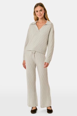 A woman wearing a light gray Cable Knots Rocio Sweater by Roller Rabbit, featuring a textured knit and V-neck, paired with matching knit pants, stands against a plain white background.