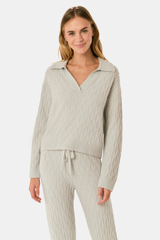 A woman wearing a light gray Cable Knots Rocio Sweater by Roller Rabbit, featuring a textured knit and V-neck, paired with matching knit pants, stands against a plain white background.