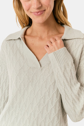 A woman wearing a light gray Cable Knots Rocio Sweater by Roller Rabbit, featuring a textured knit and V-neck, paired with matching knit pants, stands against a plain white background.
