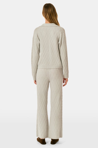 A woman wearing a light gray Cable Knots Rocio Sweater by Roller Rabbit, featuring a textured knit and V-neck, paired with matching knit pants, stands against a plain white background.