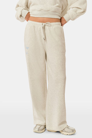 A person is wearing the Roller Rabbit Sleep Club Mimi Sweatpants in light gray, featuring wide legs and crafted from velvety soft Dream Fleece with “Sleep Club” printed in light blue on the left thigh, paired with white sneakers and a cropped white top. The background is plain white.