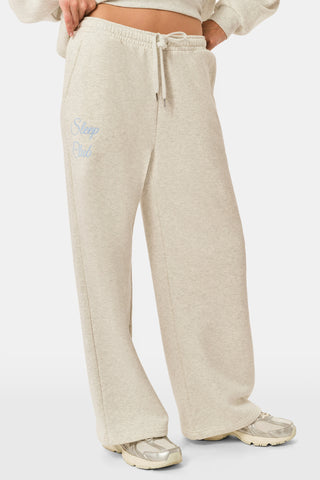 A person is wearing the Roller Rabbit Sleep Club Mimi Sweatpants in light gray, featuring wide legs and crafted from velvety soft Dream Fleece with “Sleep Club” printed in light blue on the left thigh, paired with white sneakers and a cropped white top. The background is plain white.