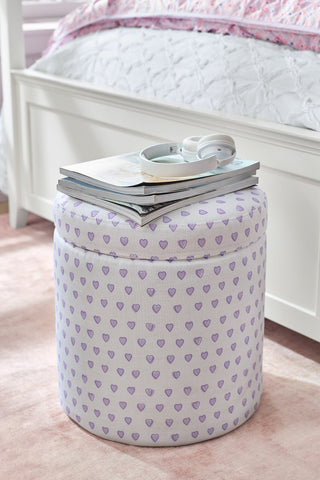 Round patterned ottoman with books on a pink carpet in a bedroom.