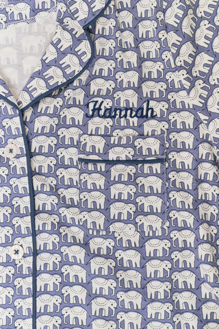 A woman wears the Hathi Polo Pajamas by Roller Rabbit, a premium cotton loungewear set with blue and white elephant patterns, including a collared button-up top and shorts, against a plain light background.