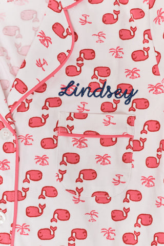 A woman wears Roller Rabbit's Moby Polo Pajamas—a women's short sleeve set with a white base and pink-red cartoon fruit prints, button-up top, and shorts. She stands against a plain light background, smiling gently.