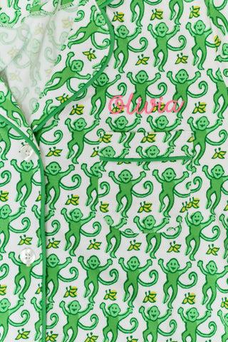 A woman wears Roller Rabbit's Monkey Polo Pajamas, a matching green and white monkey-patterned set with a button-up top and shorts. She touches her hair and smiles softly, looking slightly to the side in her stylish sleepwear.