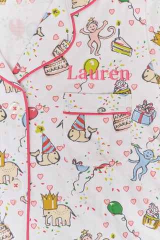 A woman smiles in the Roller Rabbit Birthday Bonanza Polo Pajamas—a women’s short set made from soft Pima cotton with colorful balloon, cake, and animal prints on white.