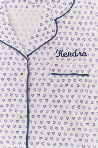A woman wears the Roller Rabbit Hearts Polo Pajamas, made from 100 cotton with a white base and small heart print. The set features a short-sleeve, button-up collared top with chest pocket and matching shorts. She stands facing forward against a plain background.