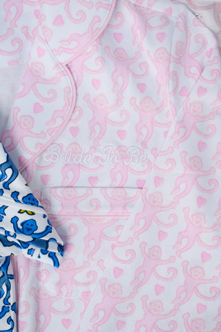 A woman with long, wavy brown hair stands barefoot against a white background, wearing the Lovely Monkey Long Sleeve Polo Pajamas in light pink with a subtle floral pattern—a comfy button-up top and matching pants perfect for relaxing nights.