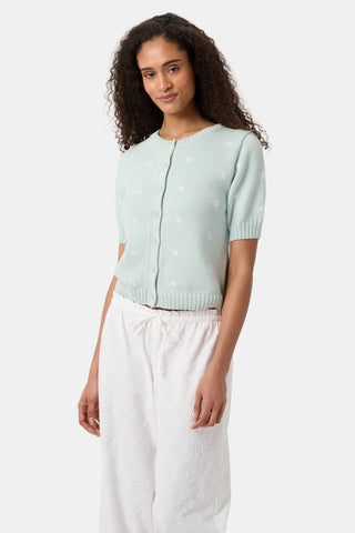 A woman with curly hair wears the Roller Rabbit Floral Bloom Nyla Cardigan, a light blue short-sleeved top with small white flowers, paired with loose white embroidered pants, standing against a plain light background.