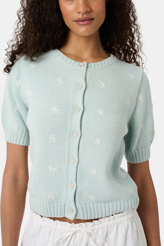 A woman with curly hair wears the Roller Rabbit Floral Bloom Nyla Cardigan, a light blue short-sleeved top with small white flowers, paired with loose white embroidered pants, standing against a plain light background.