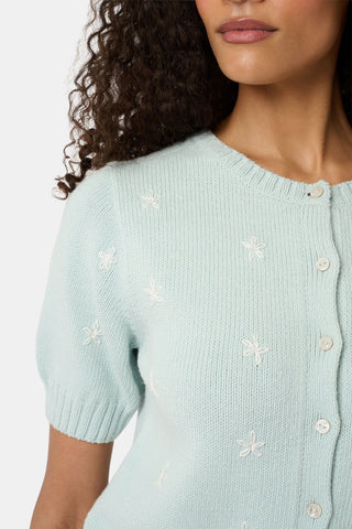 A woman with curly hair wears the Roller Rabbit Floral Bloom Nyla Cardigan, a light blue short-sleeved top with small white flowers, paired with loose white embroidered pants, standing against a plain light background.