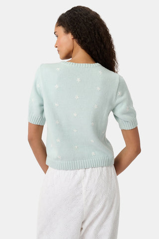 A woman with curly hair wears the Roller Rabbit Floral Bloom Nyla Cardigan, a light blue short-sleeved top with small white flowers, paired with loose white embroidered pants, standing against a plain light background.