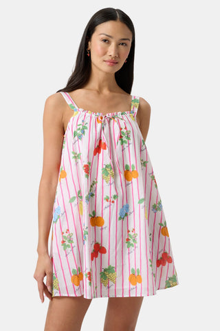A woman in the Roller Rabbit Fruits D'Ete Keaton Dress with a matching headscarf stands barefoot by a vintage white Volkswagen van, shielding her eyes from the sun. The sunny coastal backdrop highlights the dress's playful print.