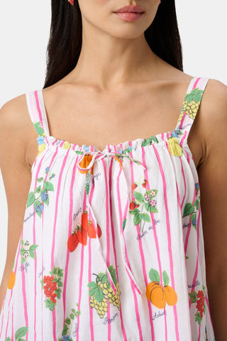 A woman in the Roller Rabbit Fruits D'Ete Keaton Dress with a matching headscarf stands barefoot by a vintage white Volkswagen van, shielding her eyes from the sun. The sunny coastal backdrop highlights the dress's playful print.
