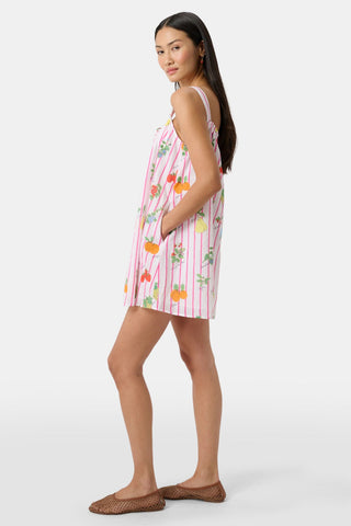 A woman in the Roller Rabbit Fruits D'Ete Keaton Dress with a matching headscarf stands barefoot by a vintage white Volkswagen van, shielding her eyes from the sun. The sunny coastal backdrop highlights the dress's playful print.