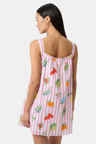 A woman in the Roller Rabbit Fruits D'Ete Keaton Dress with a matching headscarf stands barefoot by a vintage white Volkswagen van, shielding her eyes from the sun. The sunny coastal backdrop highlights the dress's playful print.