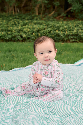 A light-skinned, dark-haired baby sits outdoors on a blue and white blanket, wearing Roller Rabbit Infant Monkey Block Stripe Zipper Footie, with green grass and bushes in the background.