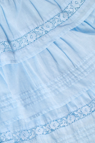 Two women stand in front of bookshelves in a brightly lit library, both wearing light blue, floral, and ruffled outfits from Roller Rabbit. One is styled in the Florence Lace Siggy Skirt, while the other wears coordinating cotton pieces. With relaxed poses and books visible behind them, they look at the camera.