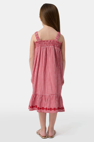 A young girl in the Roller Rabbit Kids Gingham Teagan Dress with an embroidered hem stands barefoot on a boat deck by the water, sunlight shimmering on the surface.