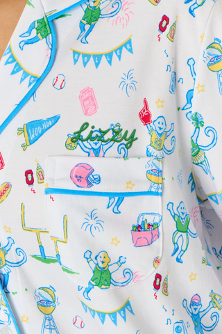 Roller Rabbit’s Game Day Long Sleeve Polo Pajamas feature a white button-up shirt and matching pants adorned with colorful, playful animal prints, festive bunting, and other cheerful designs.