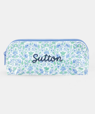 The Roller Rabbit Garden Gate Floral Makeup Bag features a rectangular zippered design with a blue and green floral pattern on a white cotton canvas background, photographed against a simple light backdrop. Perfect for organizing your beauty essentials at home or while traveling.