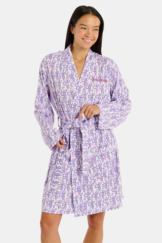 A woman smiles while wearing the TEXPIMA Get Ready With Me Blake Robe, featuring a knee-length cut and a purple and pink paisley pattern, tied at the waist, against a plain white background.